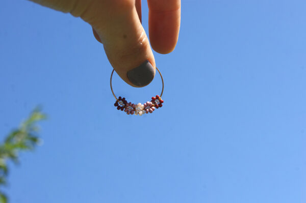 Boucle d'oreilles de type créole ornées de fleurs en perles roses tenue par une ain sur fond de ciel bleu